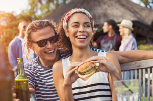 Two friends smiling at an outdoor gathering, one holding a sandwich, with people socializing in the background. Click to view a larger version.